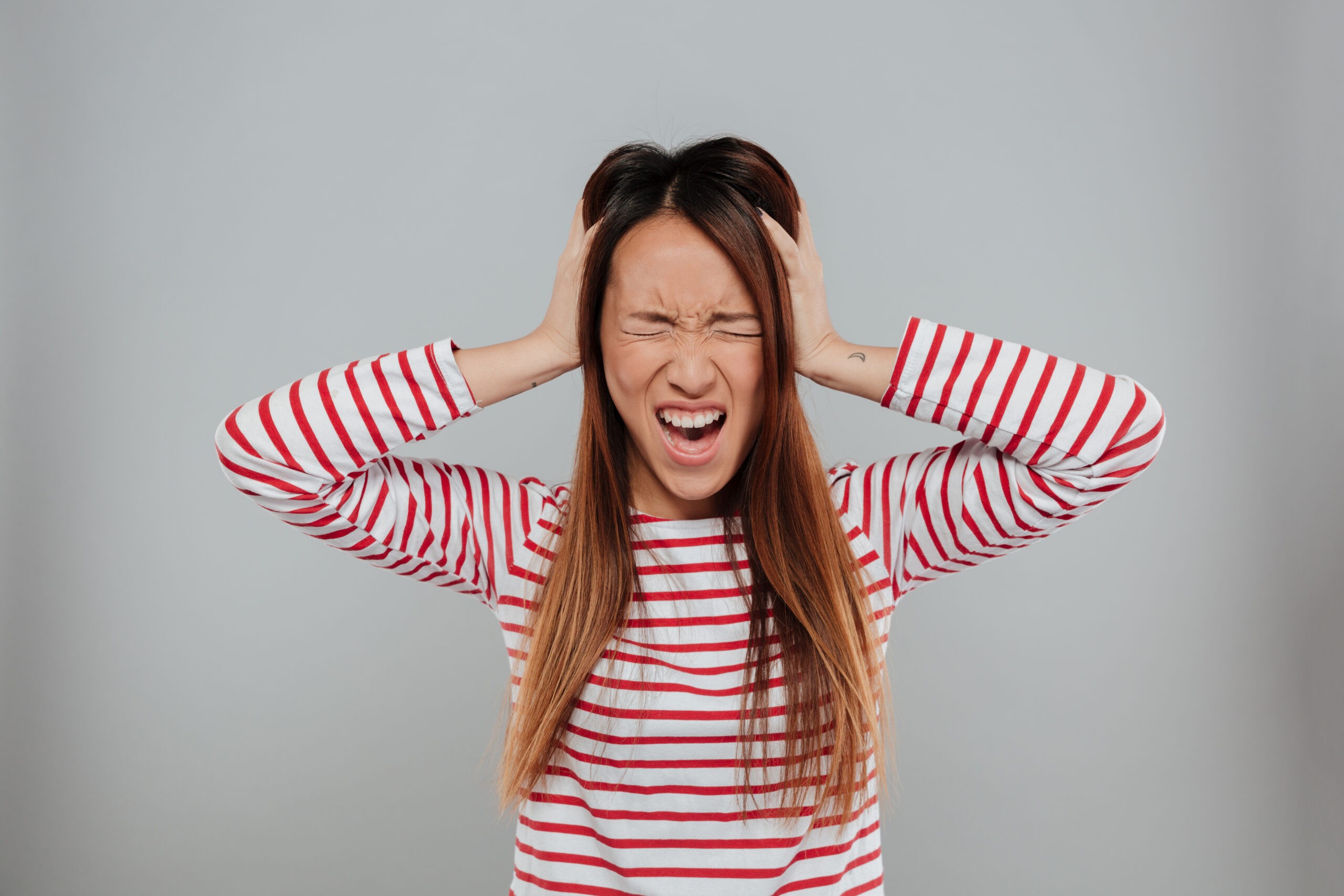 Portrait of a crazy asian girl standing with hands in her hair and screaming isolated over gray background