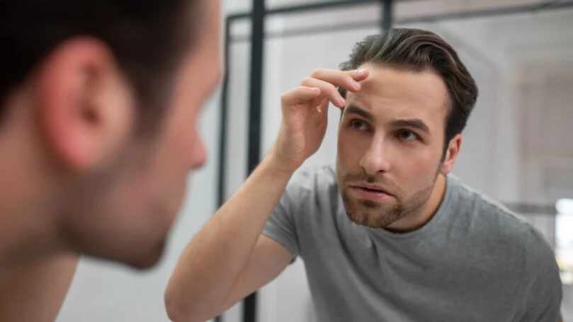 Appearance. A young man in grey tshirt looking at his reflection in the mirror