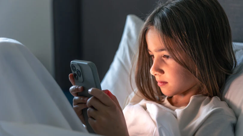 A focused girl sits among white pillows and scrolls her feed, face lit by the screen. Quiet mood, cozy home vibe and mindful browsing before sleep.